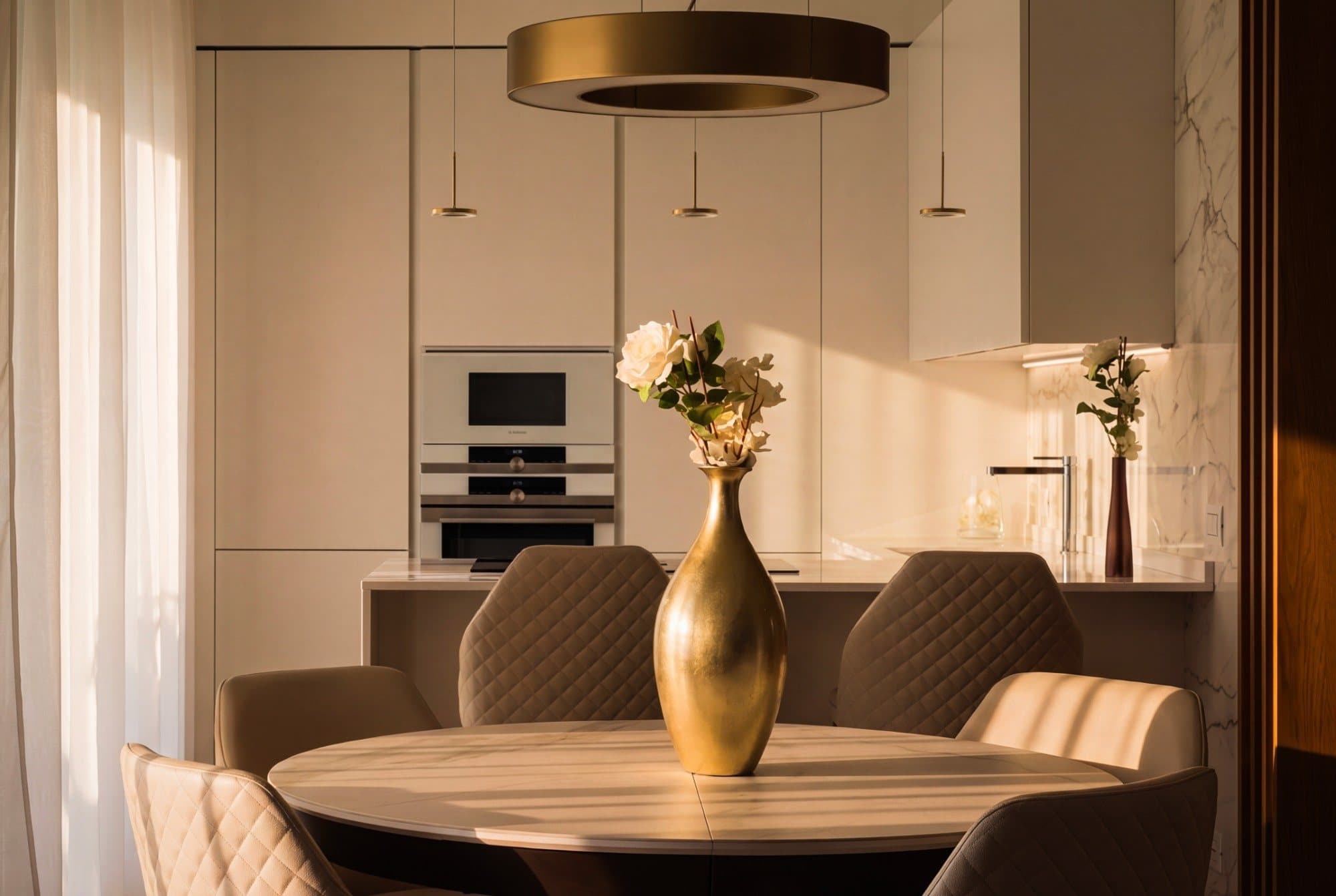 Tavolo rotondo con vaso dorato sotto una sospensione ad anello, cucina bianca sullo sfondo, progetto di Tamara Gerosa Studio a Milano